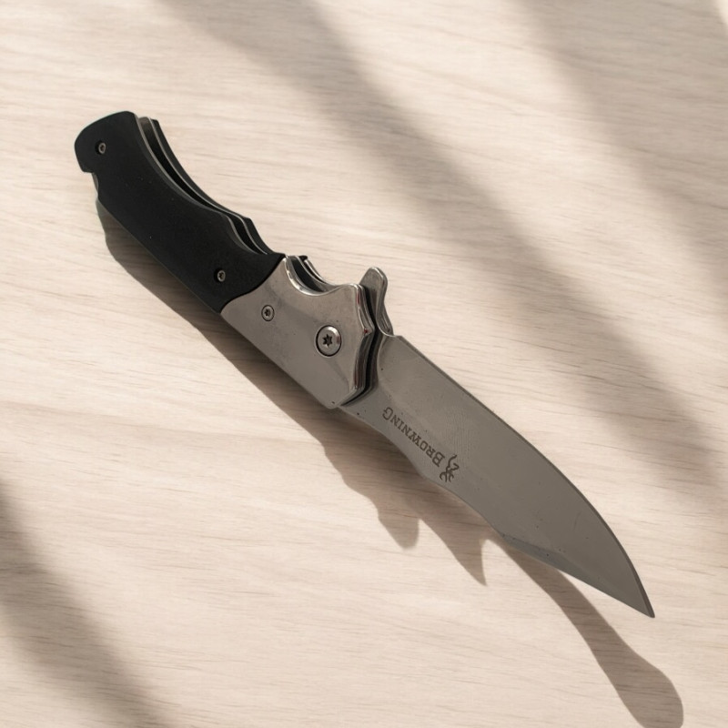 Нож Browning Silver Edge изправен върху дървена маса с отблясъци от светлина Сгъваем нож Browning със сребристо острие и черна дръжка върху светла повърхност