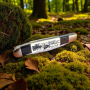 Нож с изображение на елен, разположен върху пън в зелена природа Сгъваем нож Forest Stag с гравирана дръжка, поставен върху дървена дъска и камъни